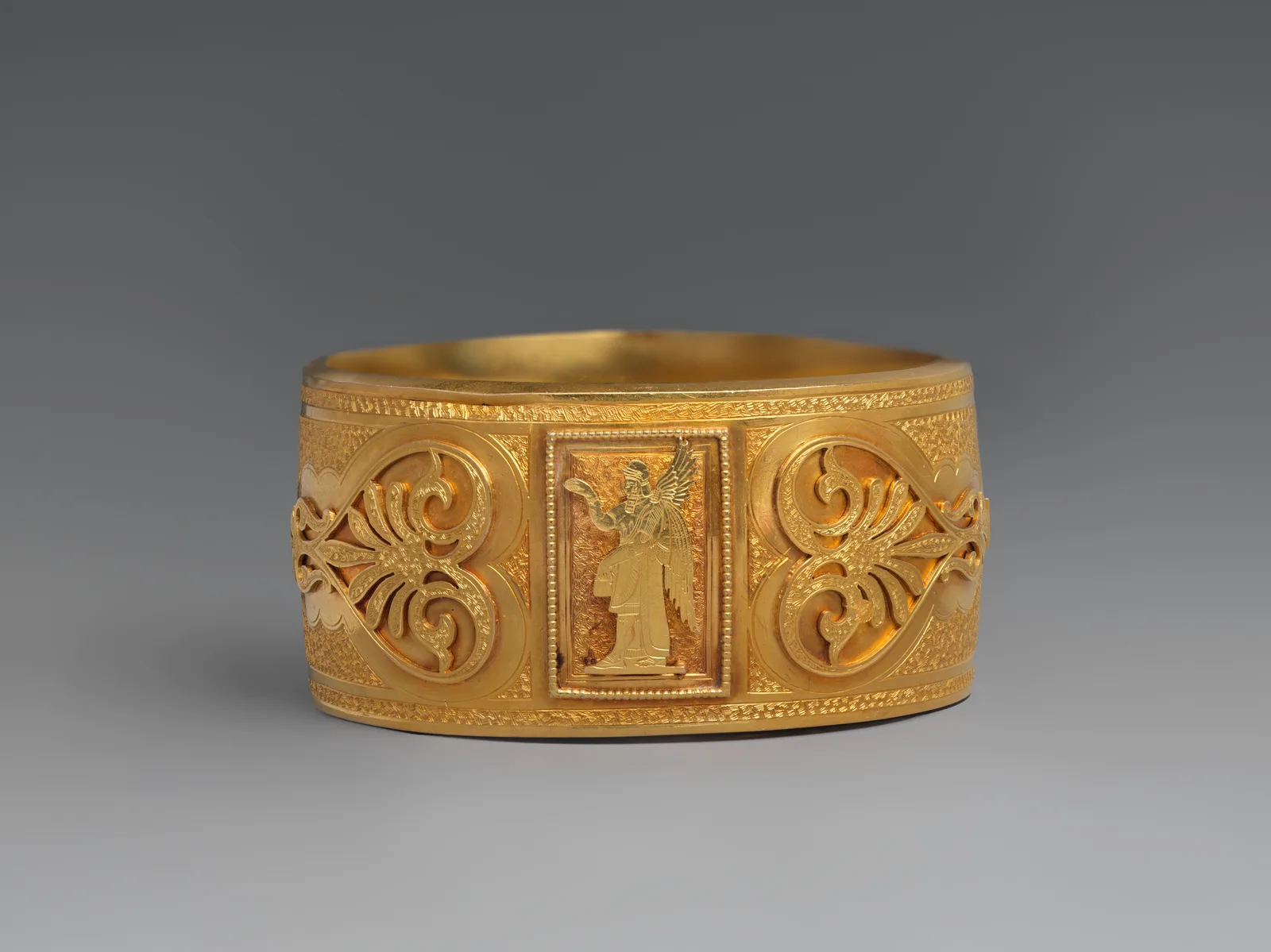 Ancha pulsera británica de oro de la década de 1870 con ornamento asirio y una figura alada en el centro
