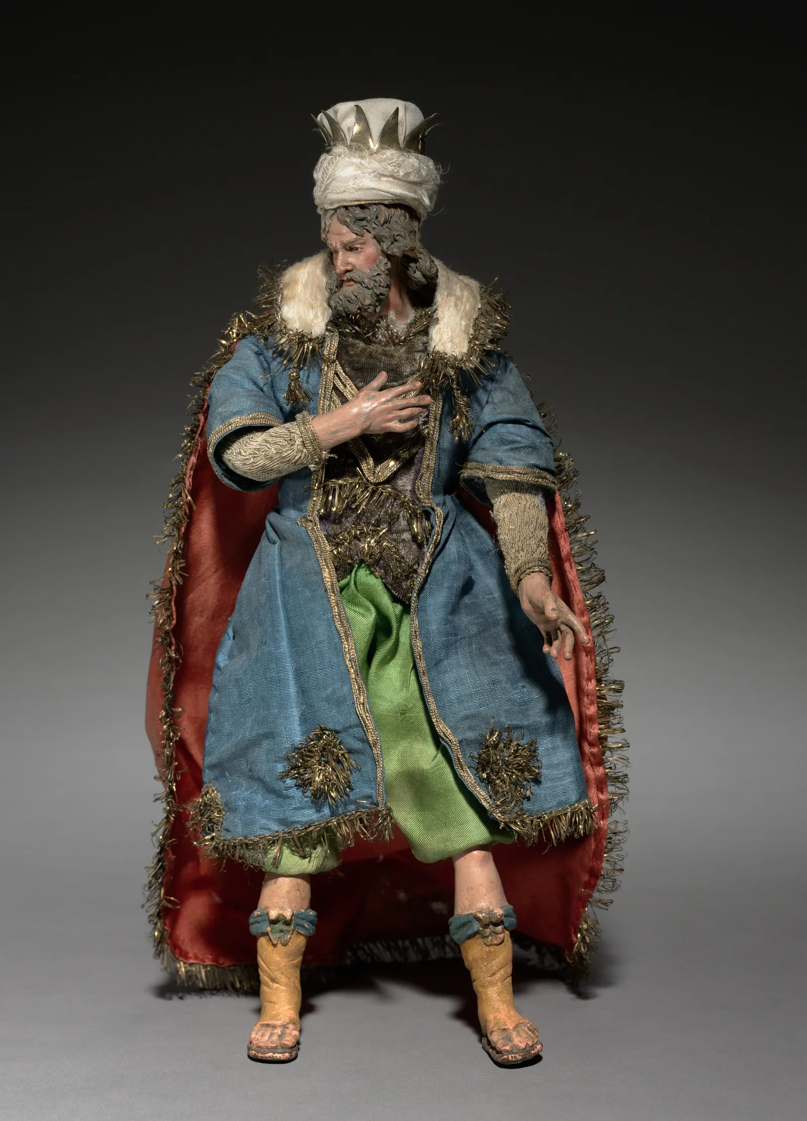 Figura de rey mago de un belén napolitano, de pie, con turbante blanco y corona, túnica azul con galones dorados y manto forrado de rojo
